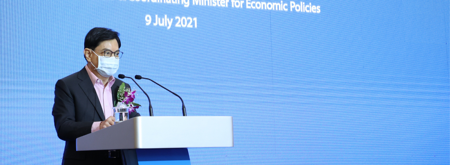 Man in suit, mask, and glasses at podium with microphones. Blue backdrop reads "9 July 2021".